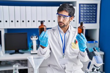 Young hispanic man scientist holding bottles at laboratory