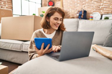 Yeni evinde dokunmatik ped ve dizüstü bilgisayar kullanan beyaz bir kadın gülümsüyor.