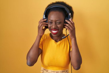 Sarı arka planda duran kıvırcık saçlı Afrikalı kadın yüksek sesli müzik sesi için kulaklık takıyor, parmaklarıyla kulaklarını kapatıyor. sağır kavramı. 