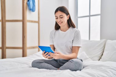 Young hispanic woman using touchpad sitting on bed at bedroom