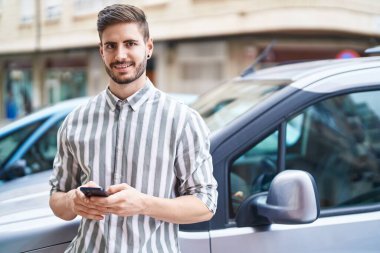 Beyaz bir adam akıllı telefon kullanıyor. Sokakta arabaya yaslanıyor.