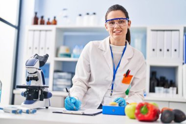 Young beautiful hispanic woman scientist weighing piece of apple writing report at laboratory