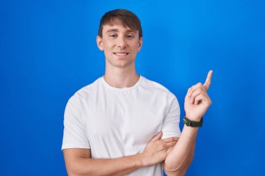 Mavi arka planda duran beyaz tenli sarışın adam, yüzünde büyük bir gülümseme, kameraya el ile işaret ediyor.. 