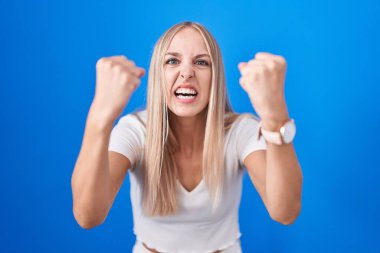 Young caucasian woman standing over blue background angry and mad raising fists frustrated and furious while shouting with anger. rage and aggressive concept. 