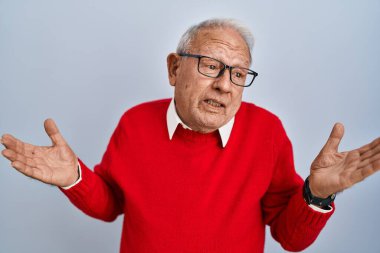 İzole edilmiş arka planda gri saçlı, kafası karışmış ve kolları ve elleri havada yaşlı bir adam. şüphe kavramı. 