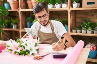 Young hispanic man florist counting dollars writing on notebook at flower shop