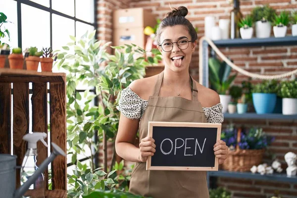 Hispanic woman working at florist holding open sign sticking tongue out happy with funny expression.
