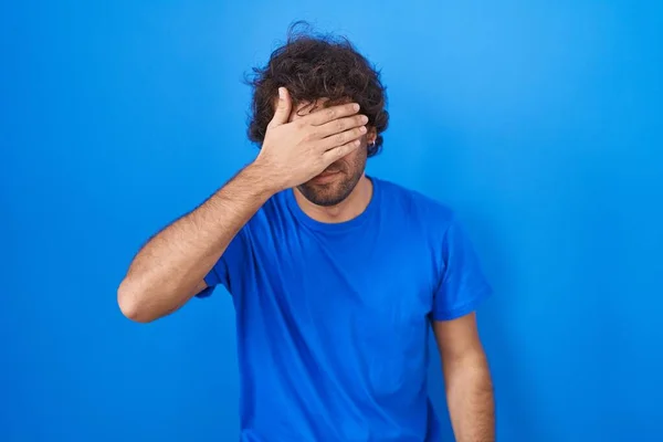 Jovem Hispânico Sobre Fundo Azul Cobrindo Olhos Com Mão Olhando — Fotografia de Stock