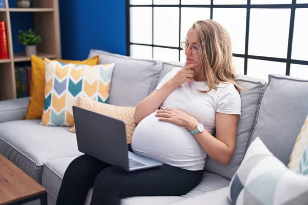 年轻的孕妇坐在家里的沙发上 用笔记本电脑认真地写着严肃的脸 手托着下巴思考着问题 思考着困惑的想法 — 图库照片