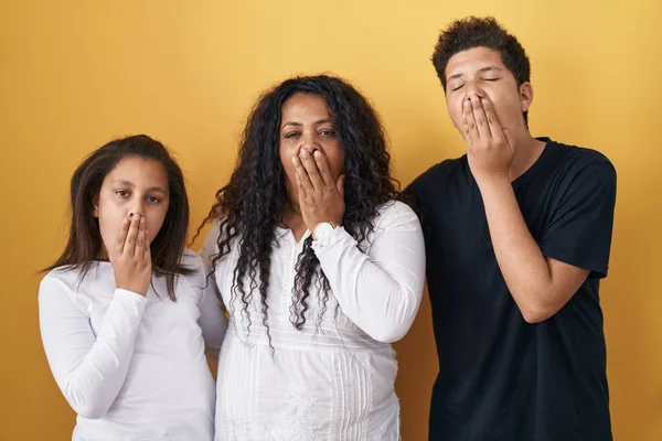 Family of mother, daughter and son standing over yellow background bored yawning tired covering mouth with hand. restless and sleepiness.