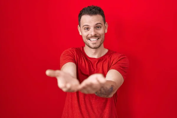 Mladý Hispánec Který Stojí Nad Červeným Pozadím Usmívá Dlaněmi Přijímá — Stock fotografie