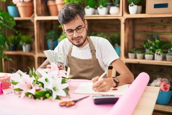 Young hispanic man florist counting dollars writing on notebook at flower shop