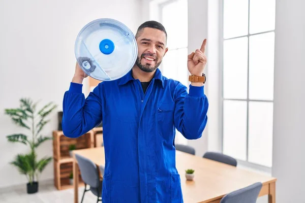 Hispanic service man holding a gallon bottle of water for delivery smiling happy pointing with hand and finger to the side