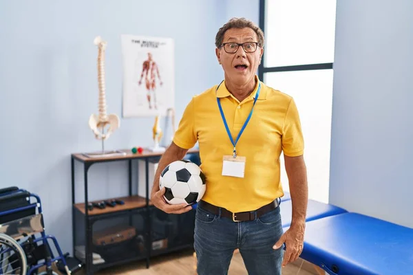 Senior man working at sport physiotherapy clinic scared and amazed with open mouth for surprise, disbelief face