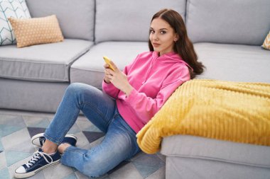 Evde yerde oturan akıllı telefonu kullanan genç bir kadın.