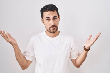Beyaz arka planda duran yakışıklı İspanyol adam. Kolları ve elleri havada, kafası karışık bir ifade. şüphe kavramı. 