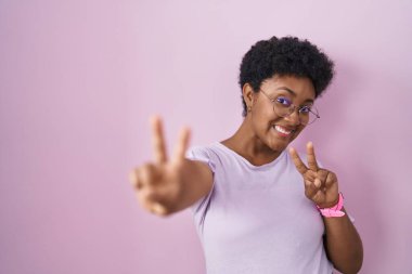 Genç Afro-Amerikan kadın pembe arka planda durup gülümsüyor kameraya bakıp zafer işareti yapıyor. İki numara.. 