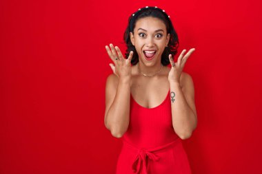 Young hispanic woman standing over red background celebrating crazy and amazed for success with arms raised and open eyes screaming excited. winner concept 
