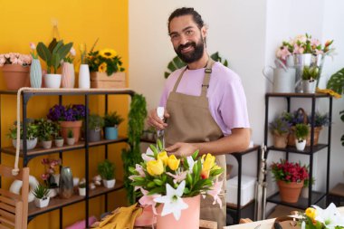 Young hispanic man florist using diffuser watering flowers at flower shop