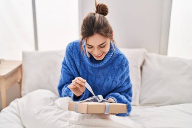 Young beautiful hispanic woman unpacking gift sitting on bed at bedroom