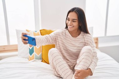 Genç ve güzel İspanyol kadın, yatak odasında oturmuş akıllı telefonuyla selfie çekiyor.