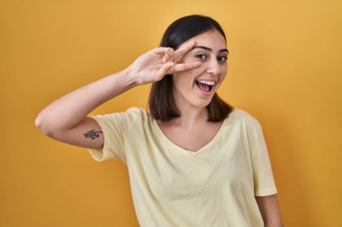 Hispanic girl wearing casual t shirt over yellow background doing peace symbol with fingers over face, smiling cheerful showing victory 