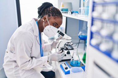 African american woman wearing scientist uniform and medical mask using microscope at laboratory