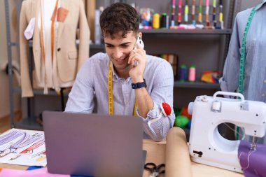 Young hispanic man tailor talking on smartphone using laptop at atelier