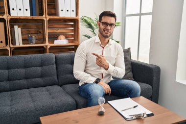 Young hispanic man with beard working at consultation office pointing aside worried and nervous with forefinger, concerned and surprised expression 