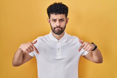 Sarı arka plan üzerinde duran Arap adam üzgün ve üzgün görünüyor, parmaklarıyla yön gösteriyor, mutsuz ve depresif.. 