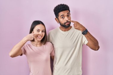 Young hispanic couple together over pink background pointing with hand finger to face and nose, smiling cheerful. beauty concept 