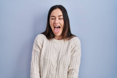 Young brunette woman standing over blue background sticking tongue out happy with funny expression. emotion concept. 