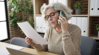Middle age woman with grey hair business worker talking on smartphone reading document at office