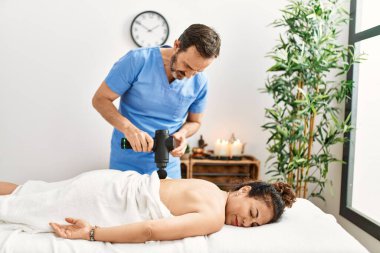 Orta yaşlı erkek ve kadın terapist üniforması giyiyorlar. Güzellik merkezinde perküsyon tabancası kullanıyorlar.