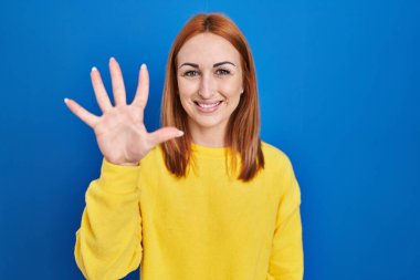 Young woman standing over blue background showing and pointing up with fingers number five while smiling confident and happy. 