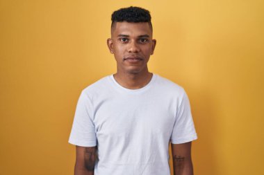 Young hispanic man standing over yellow background relaxed with serious expression on face. simple and natural looking at the camera. 