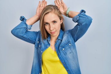 Young blonde woman standing over blue background doing bunny ears gesture with hands palms looking cynical and skeptical. easter rabbit concept. 