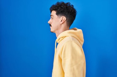 Hispanic man standing over blue background looking to side, relax profile pose with natural face and confident smile. 