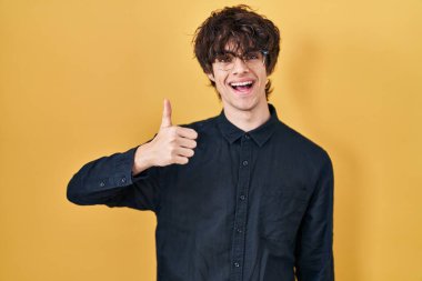 Young man wearing glasses over yellow background doing happy thumbs up gesture with hand. approving expression looking at the camera showing success. 