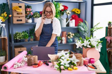 Young blonde woman florist using laptop talking on smartphone at florist shop