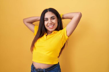Young arab woman standing over yellow background relaxing and stretching, arms and hands behind head and neck smiling happy 