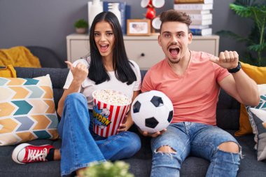 Genç İspanyol çift futbol holiganları top tutuyorlar ve patlamış mısır yiyorlar başparmağıyla yan tarafa doğru gülümsüyorlar. 