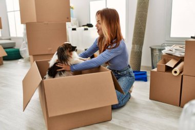 Beyaz bir kadın yeni evinde elinde karton kutunun köpeğiyle yerde oturuyor.