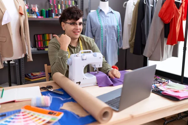 Young non binary man dressmaker designer on video call with laptop pointing thumb up to the side smiling happy with open mouth