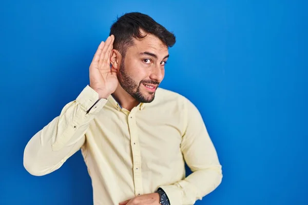 噂やゴシップに耳を傾ける耳の上に手で笑みを浮かべて青い背景に立ってハンサムなヒスパニック系の男 聴覚障害の概念 — ストック写真