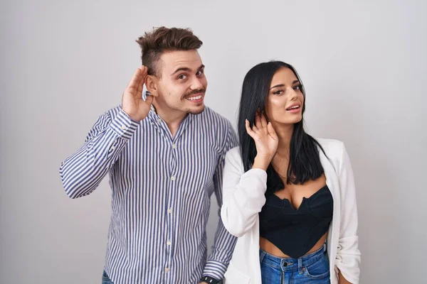 若いヒスパニック系のカップルの白い背景に手を耳に噂やゴシップに耳を傾け笑みを浮かべて立っている 聴覚障害の概念 — ストック写真