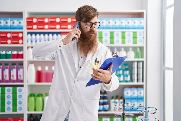 Young Redhead Man Pharmacist Talking Smartphone Reading Document Pharmacy — стоковое фото