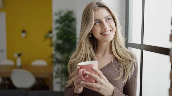 Młoda Blondynka Uśmiecha Się Pewnie Pijąc Kawę Domu — Zdjęcie stockowe