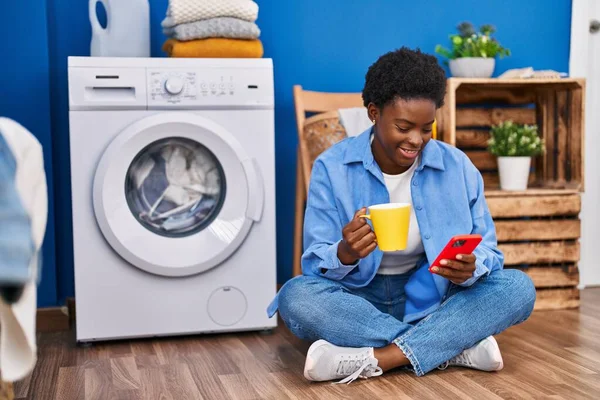 非洲裔美国妇女在洗衣房用智能手机和咖啡等洗衣机 — 图库照片