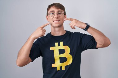 Bitcoin tişörtü giyen, neşeli gülümseyen ve parmaklarıyla ve ağzıyla işaret eden beyaz sarışın bir adam. diş sağlığı kavramı. 
